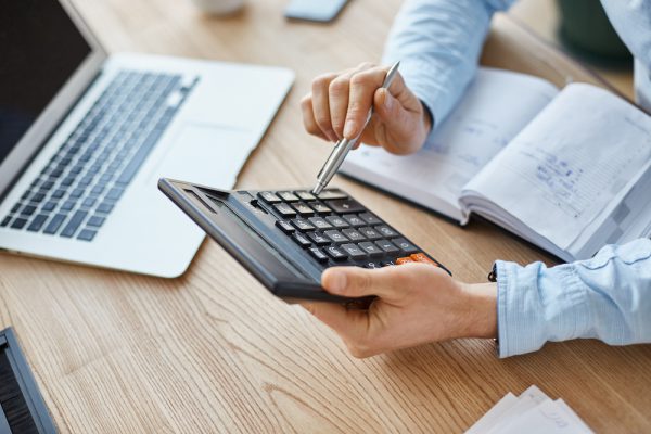 Cropped view of professional serious finance manager, holding calculator in hands, checking company month's profits, looking through details on laptop, writing down information for meeting with director